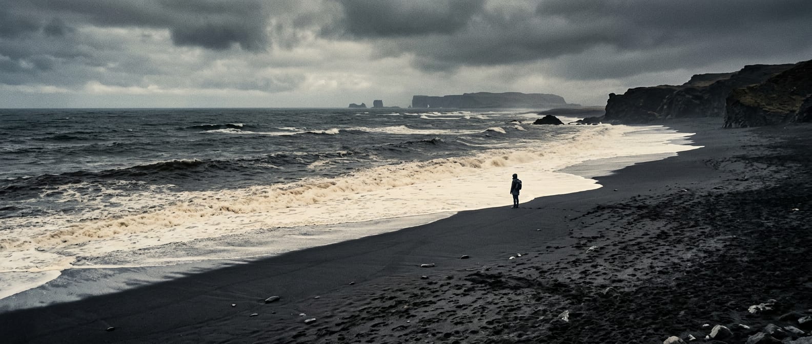 A lone figure on a black volcanic beach beside pale surf under a heavy grey sky