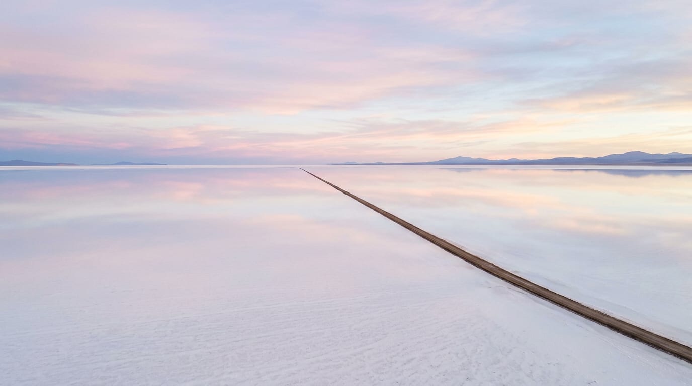 A vast white salt flat at dawn under a pink and lavender sky, with one straight dirt road running into the distance