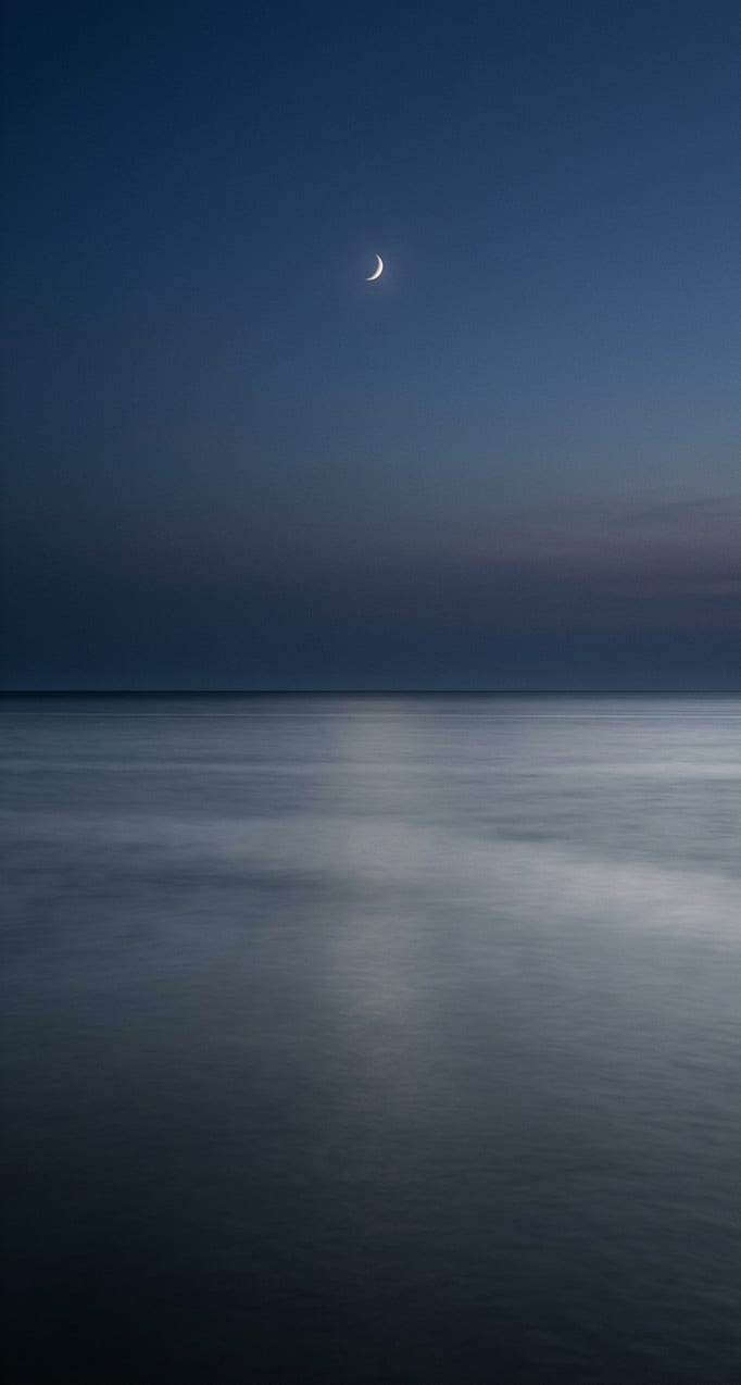 A calm sea at dusk under a deep indigo sky with a thin crescent moon